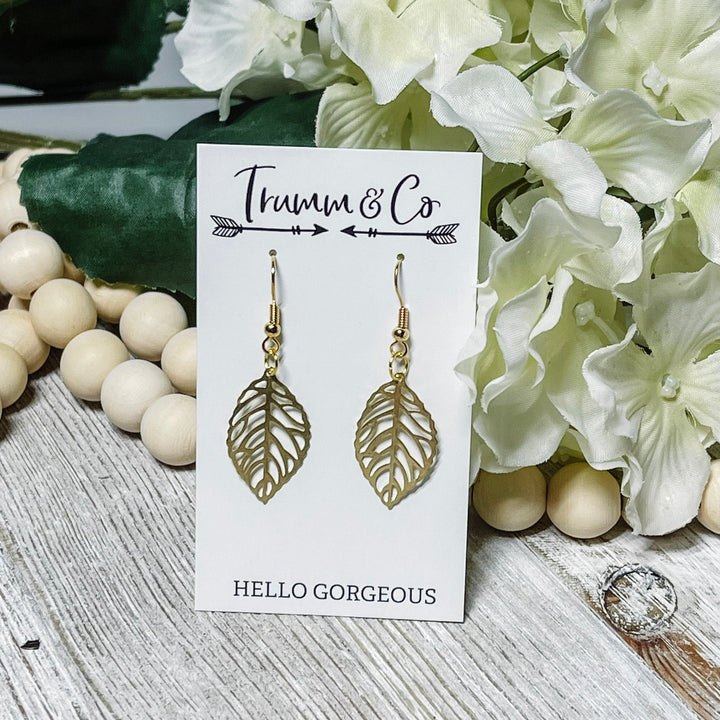 Intricate Brass leaf dangle earrings on white Trumm & Co earring card leaning on cream colored flowers